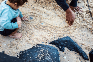 A toddler crouched on the ground as adult hands draw  pictures in the sand. Source: Destination Gold Coast